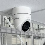 Hikvision AI-powered CCTV camera mounted on a ceiling inside a secure storage facility, overlooking mesh security fencing.